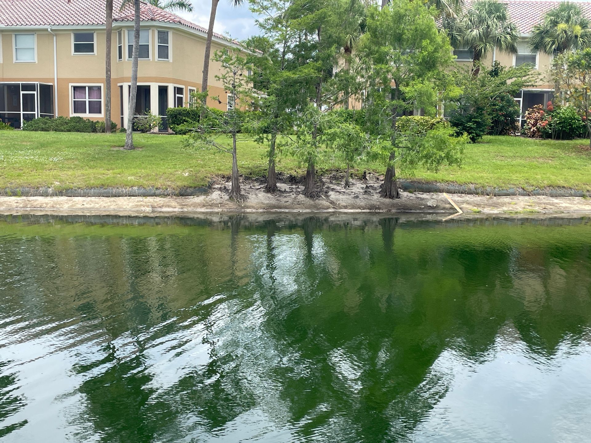 Canal with calm water reflecting trees, grassy bank, and tan homes across the shore