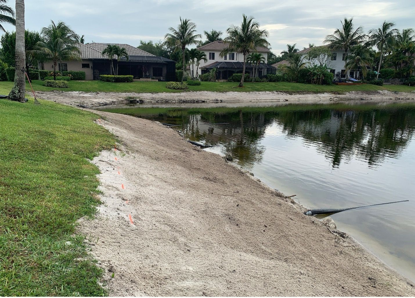 Lakeside view with houses, lush trees, and sand-covered shoreline. Overcast sky reflecting in the water.
