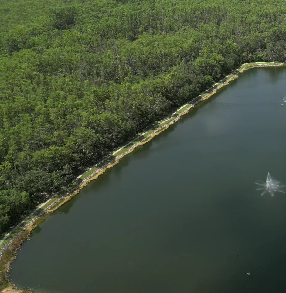 Aerial view of a dark lake bordered by a green forest with a small walking path. Water fountains visible.