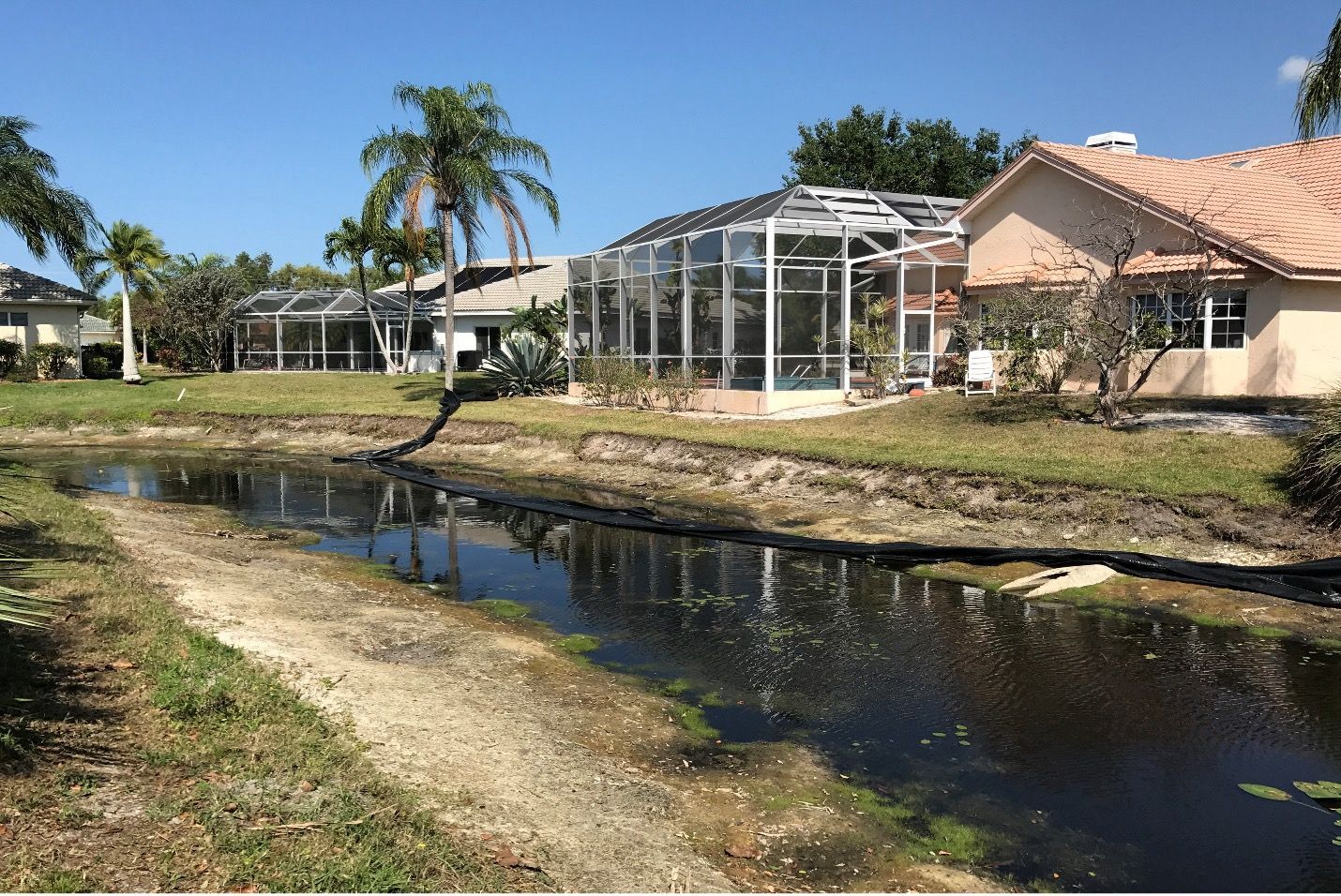 Homes with screened patios line a murky canal with patchy grass on a sunny day.