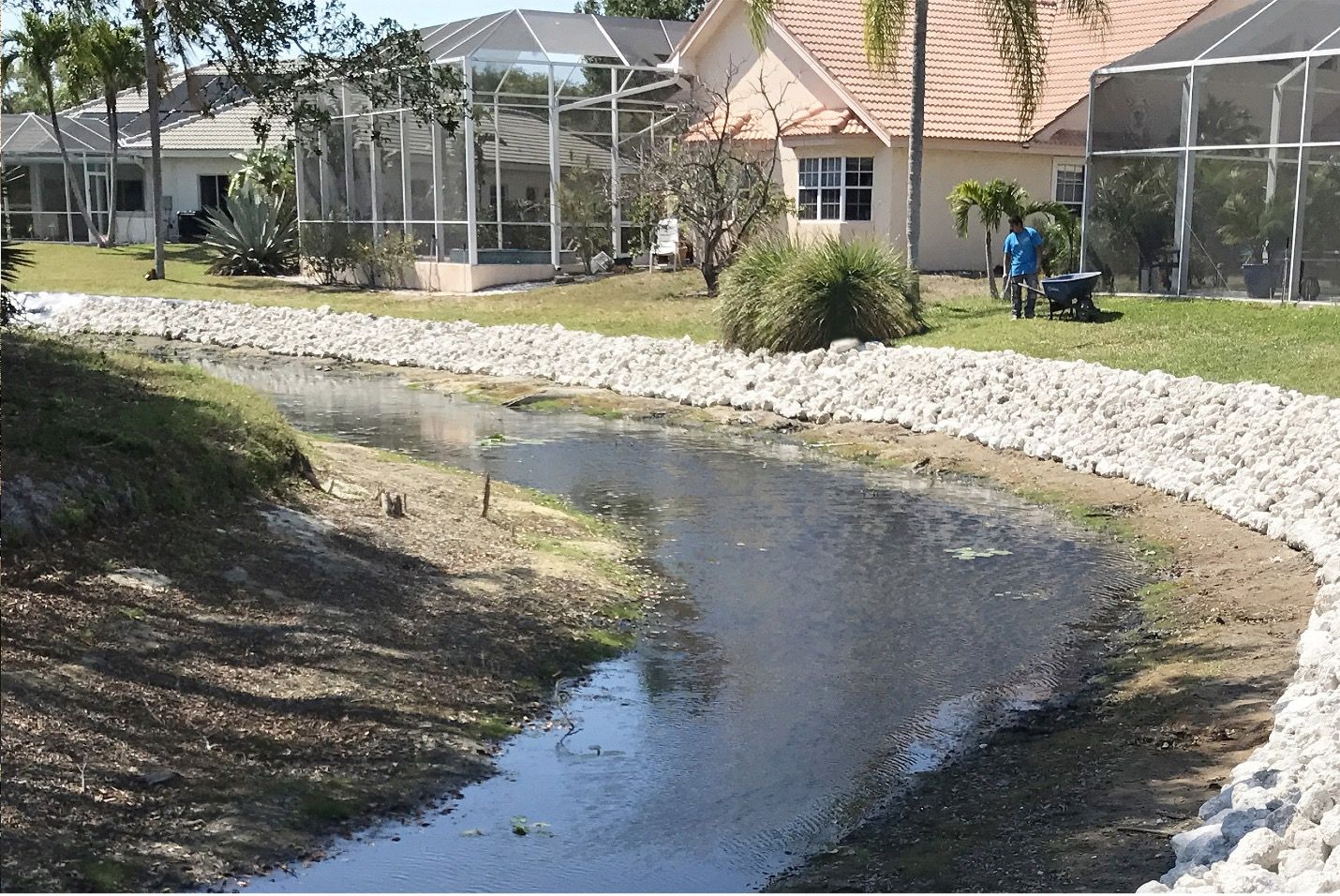 A residential waterway with rock-lined banks. Houses and a person working in the background.