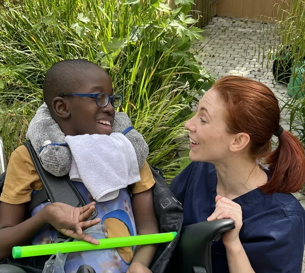 Smiling adult in blue scrubs holding a smiling child with glasses in a wheelchair outdoors
