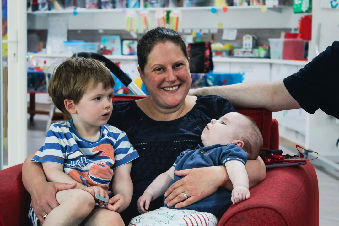 Smiling woman holding a baby beside a young child in a red chair, indoors