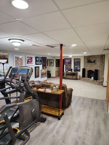 A basement features a workout area with an elliptical, a brown sofa, a red support pole, and a stone fireplace.
