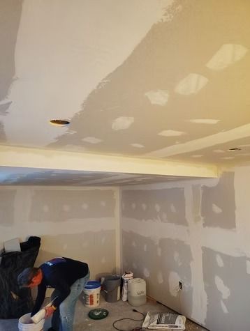 A person in a dark shirt works on patching and smoothing drywall on the walls and ceiling of an unfinished room.