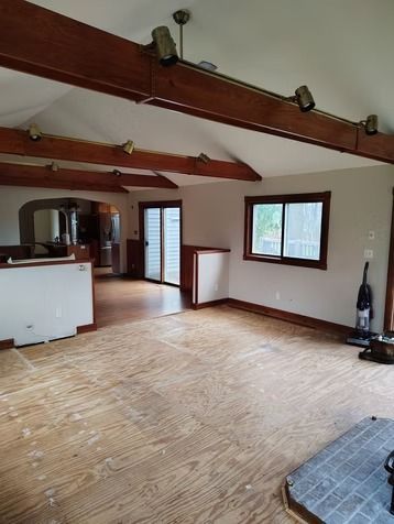 A room with exposed wood ceiling beams, wood subflooring, light walls, a brick hearth, and a doorway.