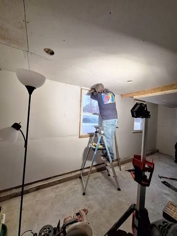 A person stands on a step ladder to work on a window frame in a room under construction.