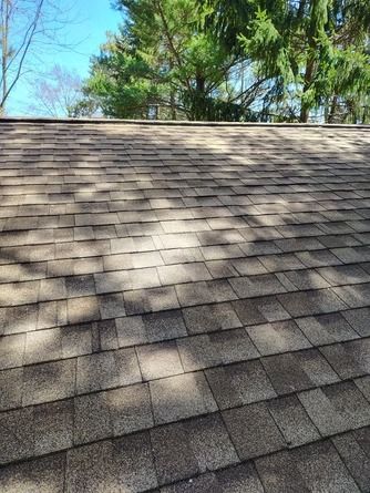 Close-up view of an asphalt shingle roof in shades of brown, with trees and a clear blue sky in the background.