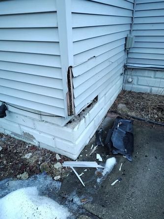 Damaged light gray house siding at the corner, with missing panels and debris scattered on the ground.
