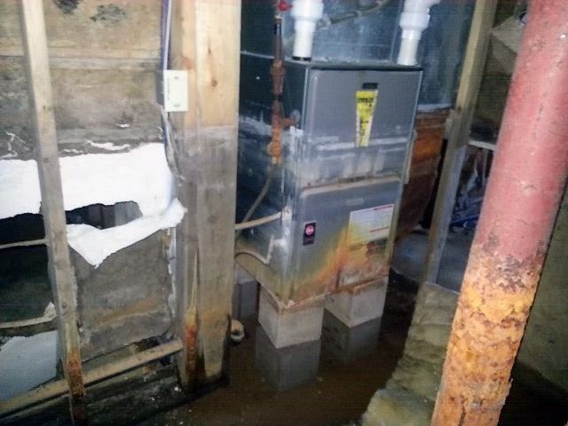 A furnace standing on concrete blocks in a damp basement with rusted floor joists.