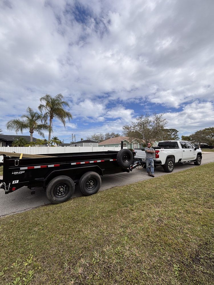 A person standing by a white truck towing a black dump trailer on a grassy street under a cloudy sky.