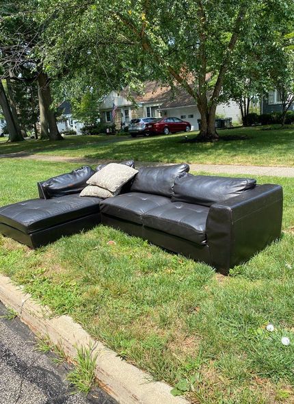 Black leather sectional sofa discarded on a grassy lawn near a curb.