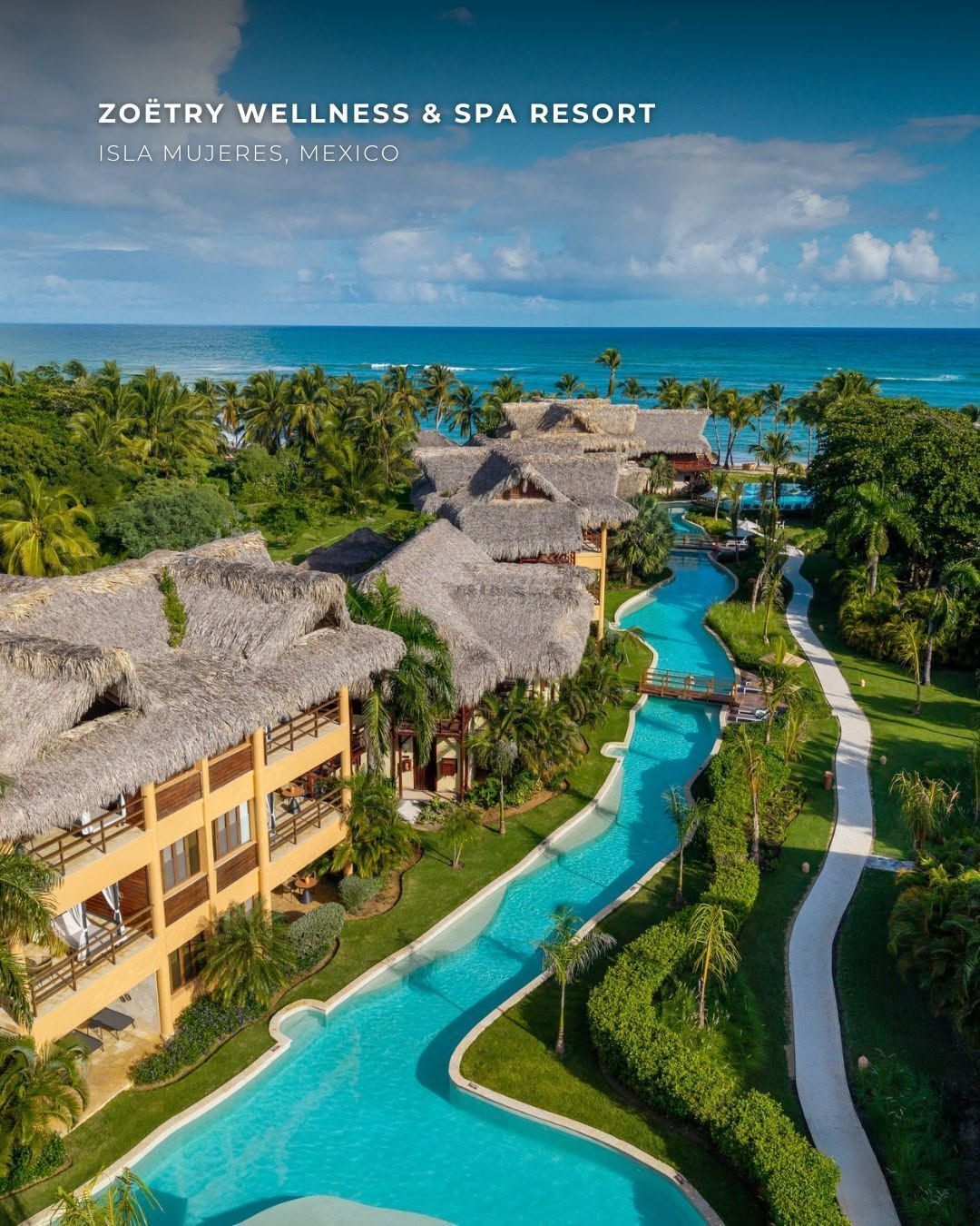 Aerial view of a tropical resort with thatched-roof buildings and a winding turquoise pool by the ocean.