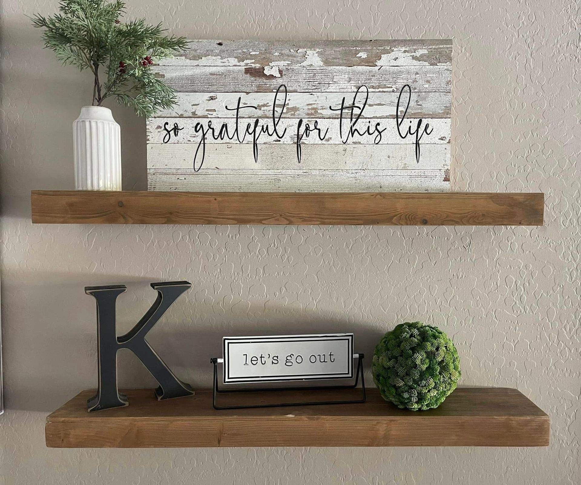 Two wooden shelves on a textured wall; the top one has a vase and sign that says 