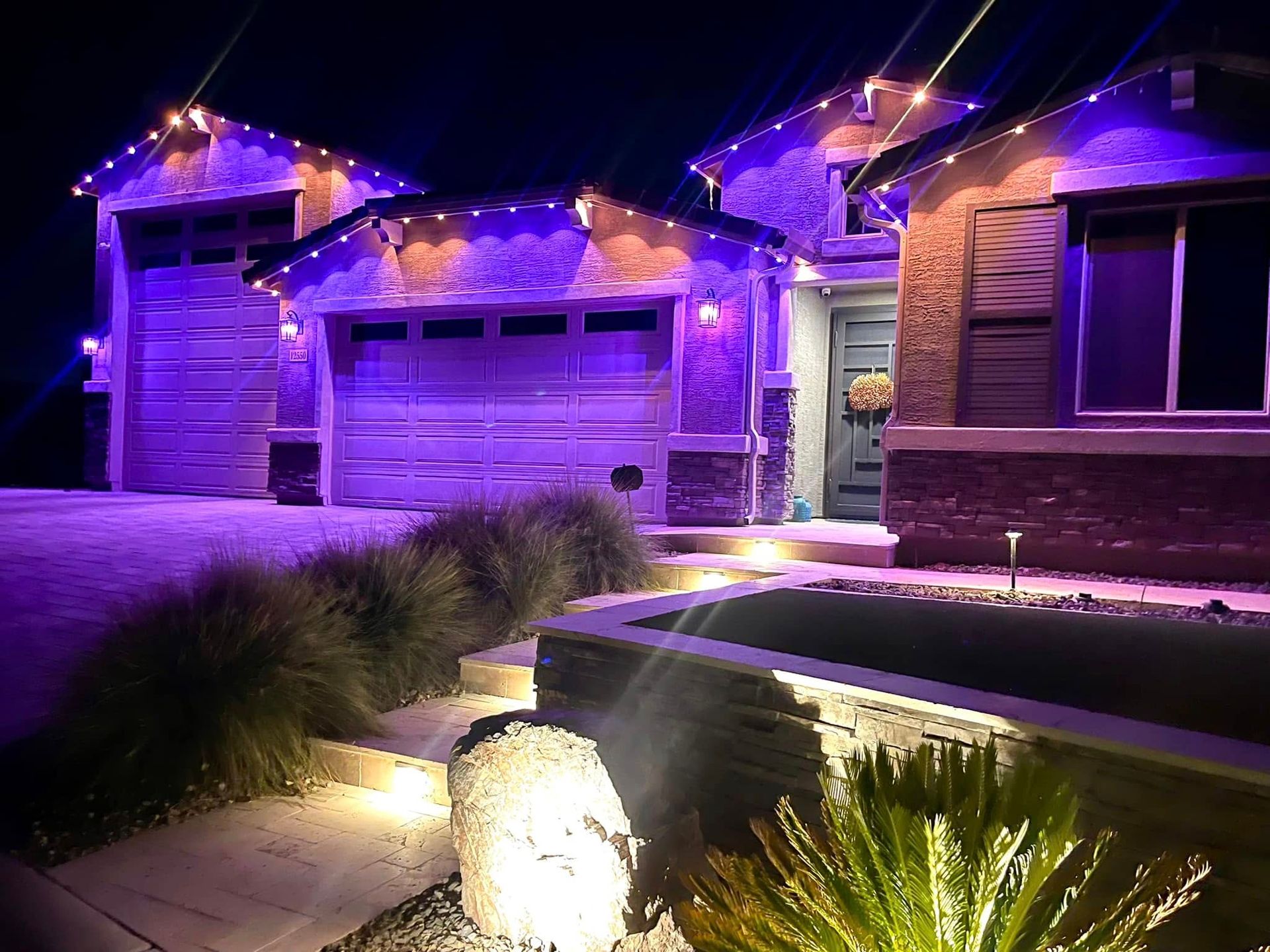 A house at night with purple lighting, decorated with string lights, and lit landscaping.