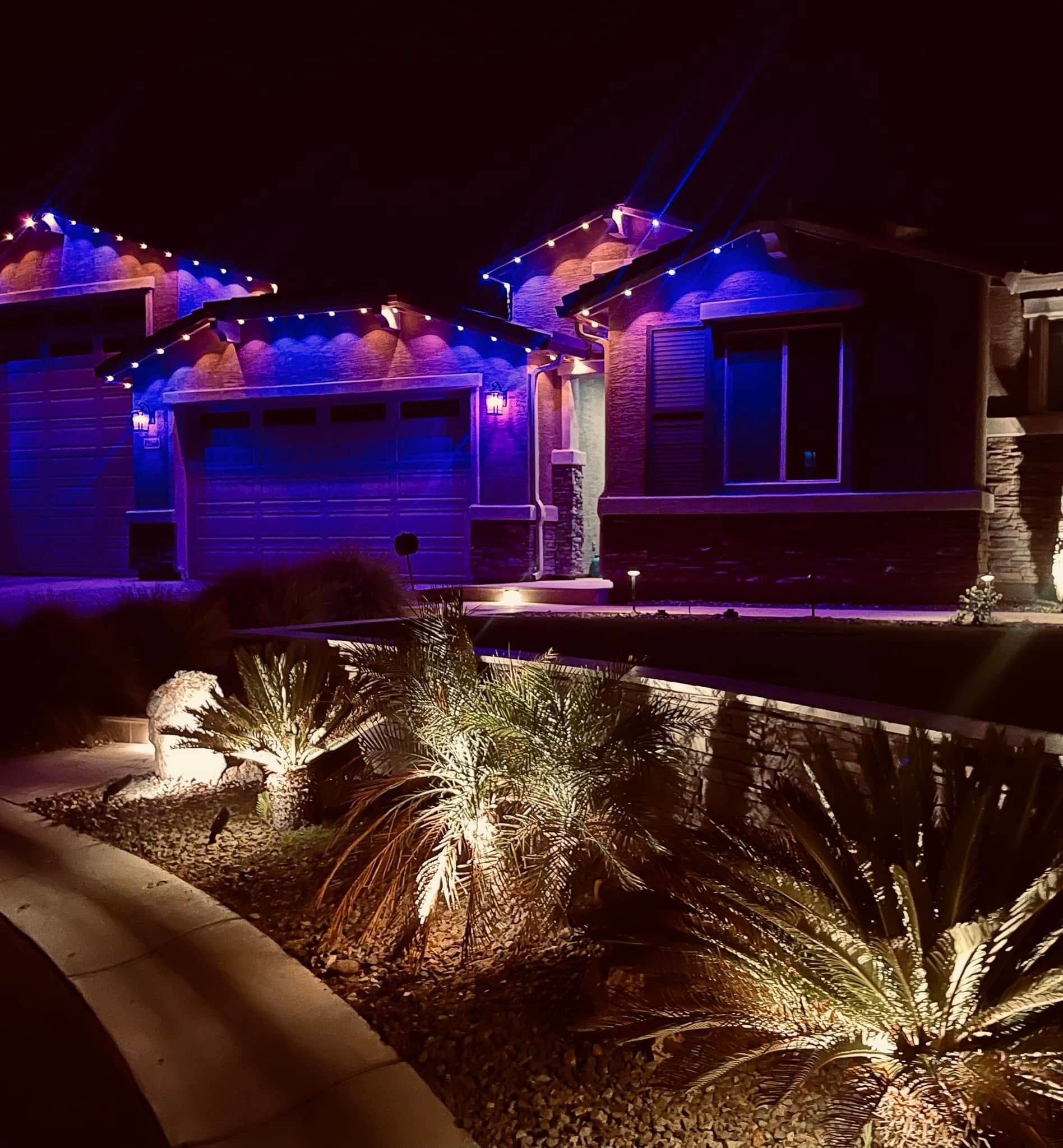 House exterior at night, lit with blue and orange lights; landscaping features lit with spotlights.