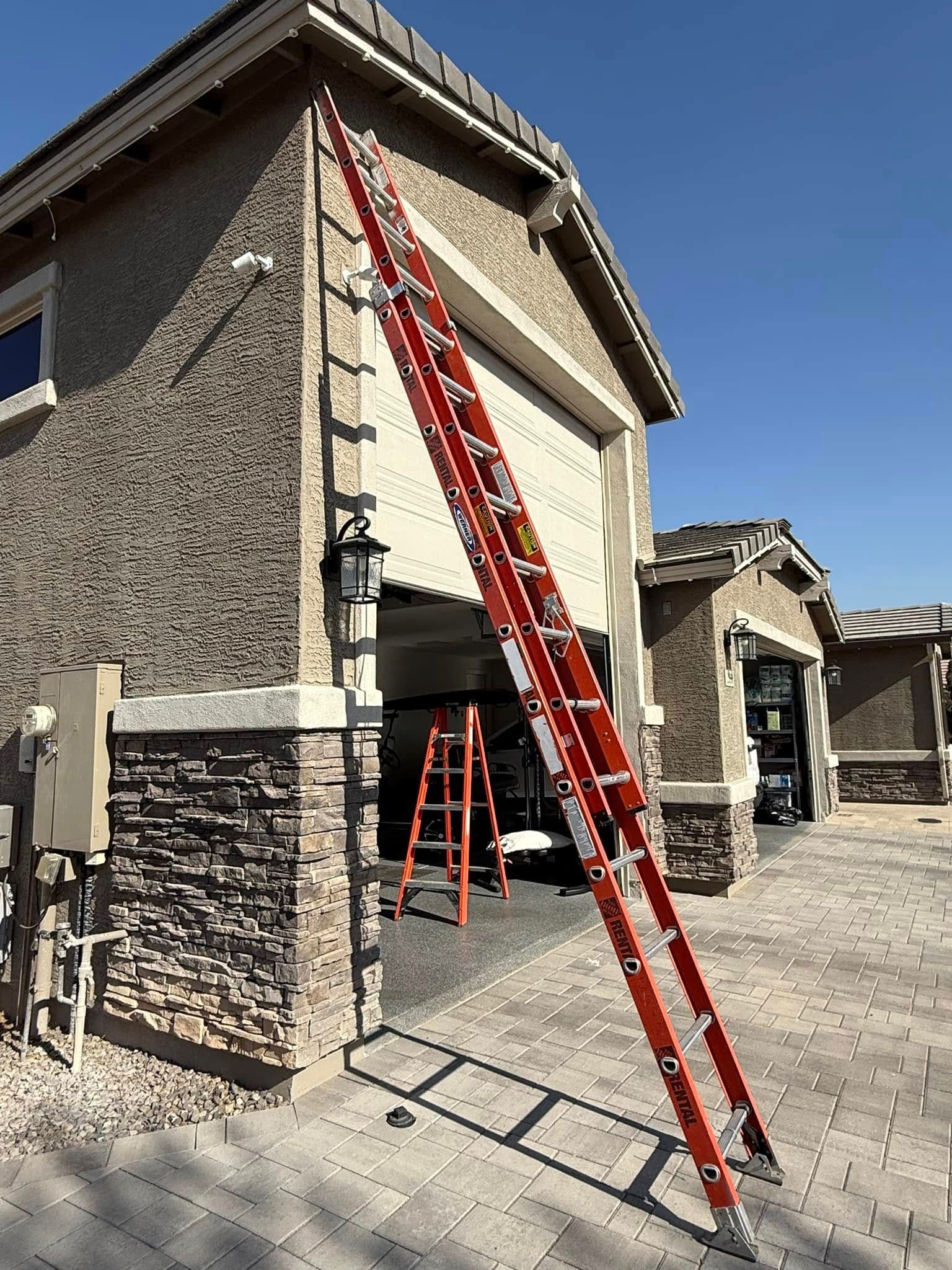 Red ladder leaning against a stucco building with open garage door and smaller ladder inside.