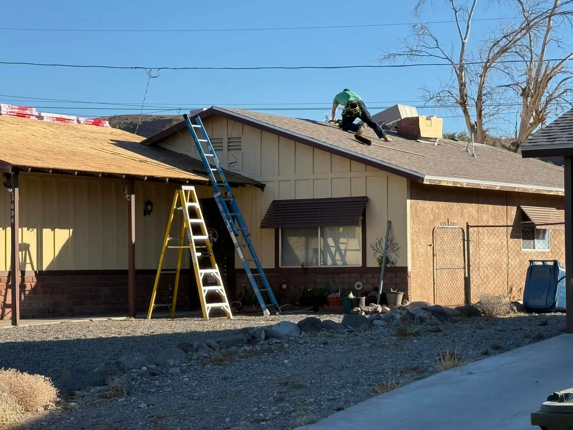 Person on a roof installing roofing. Ladders and materials are present. Building is yellow and brick.