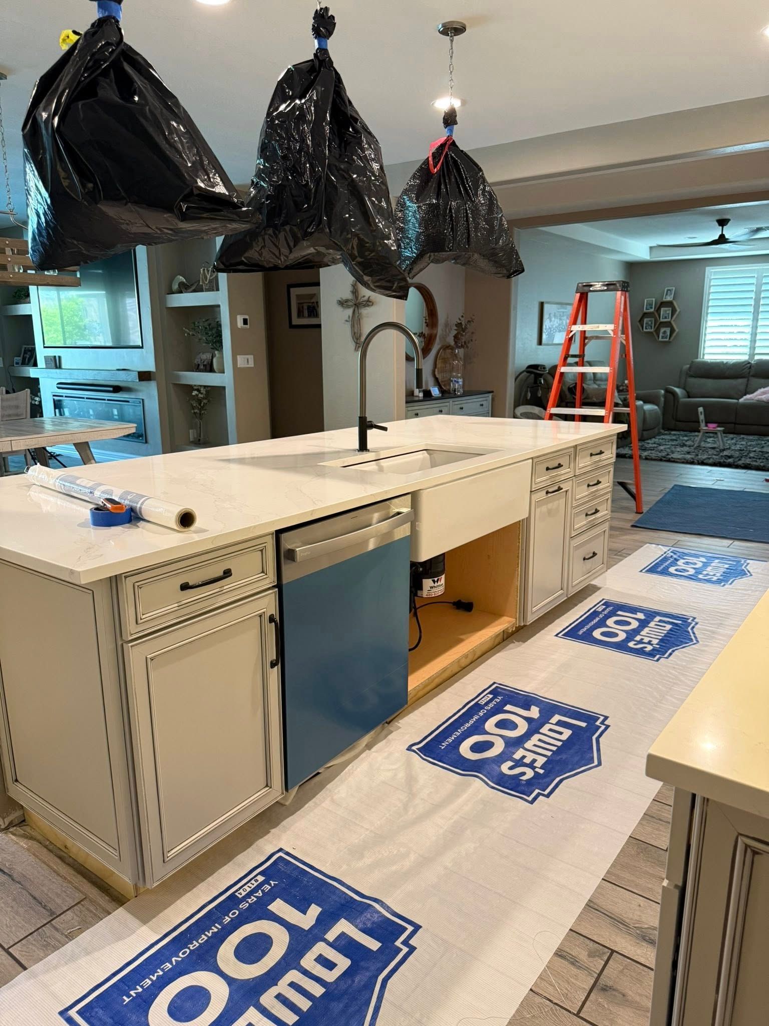 Kitchen renovation in progress, island with new sink, dishwasher, covered floors, black bags hanging from ceiling.