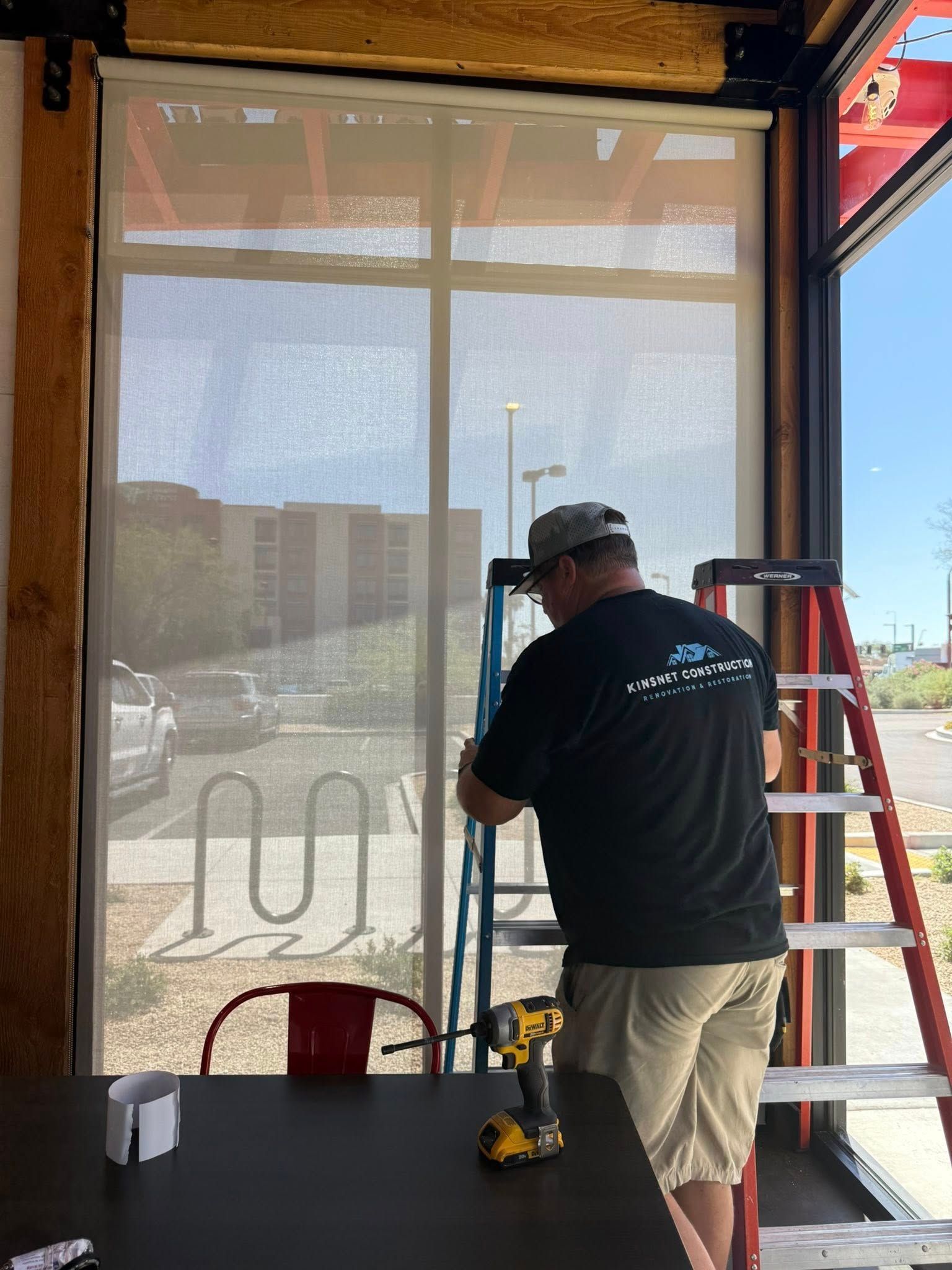 Man installing a roller shade in a building. He is on a ladder with a power drill.