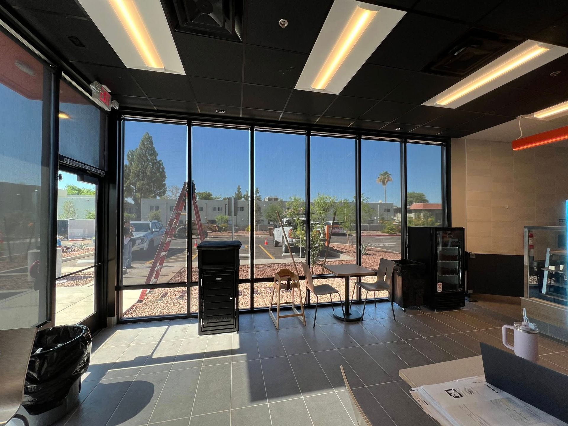 Interior shot with large windows overlooking a city view, table and chairs inside, black ceiling, sunny day.