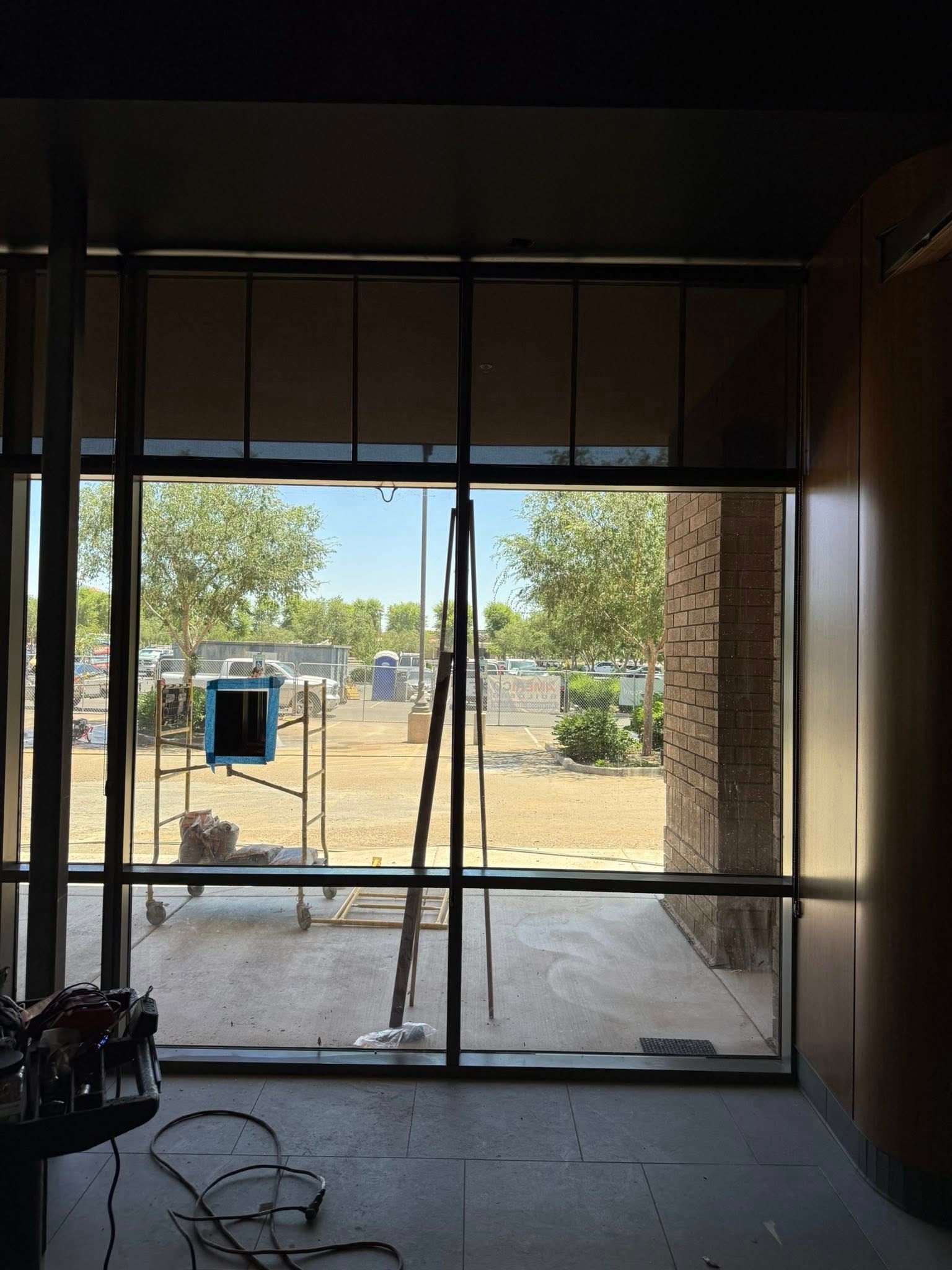 View through a large window. Construction site visible with scaffolding and trees. Interior shows unfinished space.