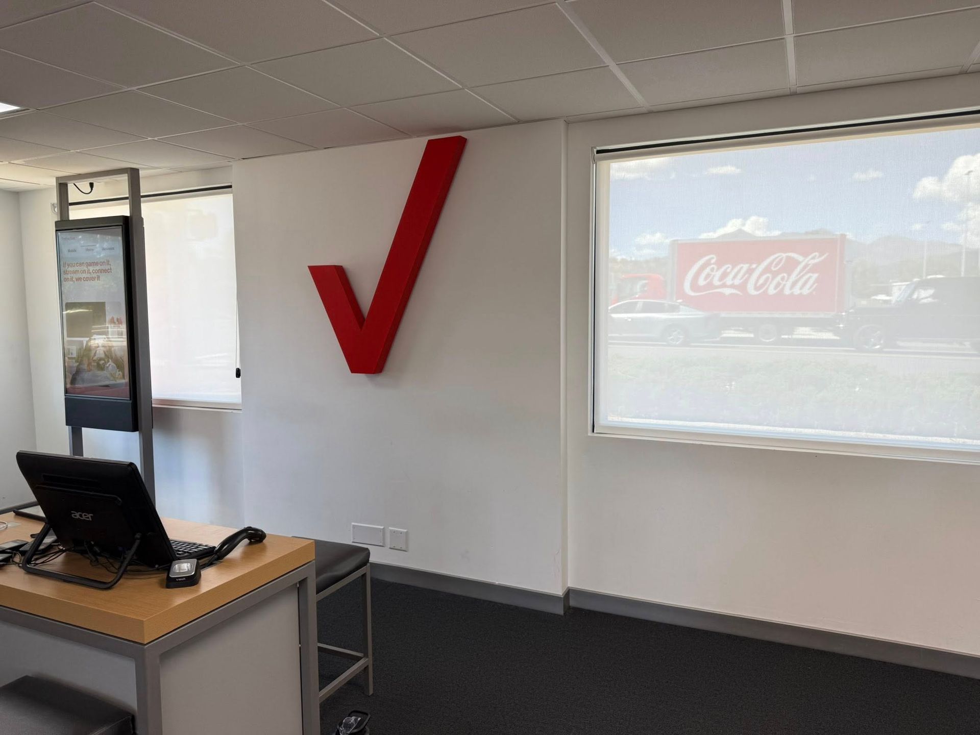 Verizon office with a large red checkmark logo on white wall, window with a Coca-Cola sign outside, and desk.