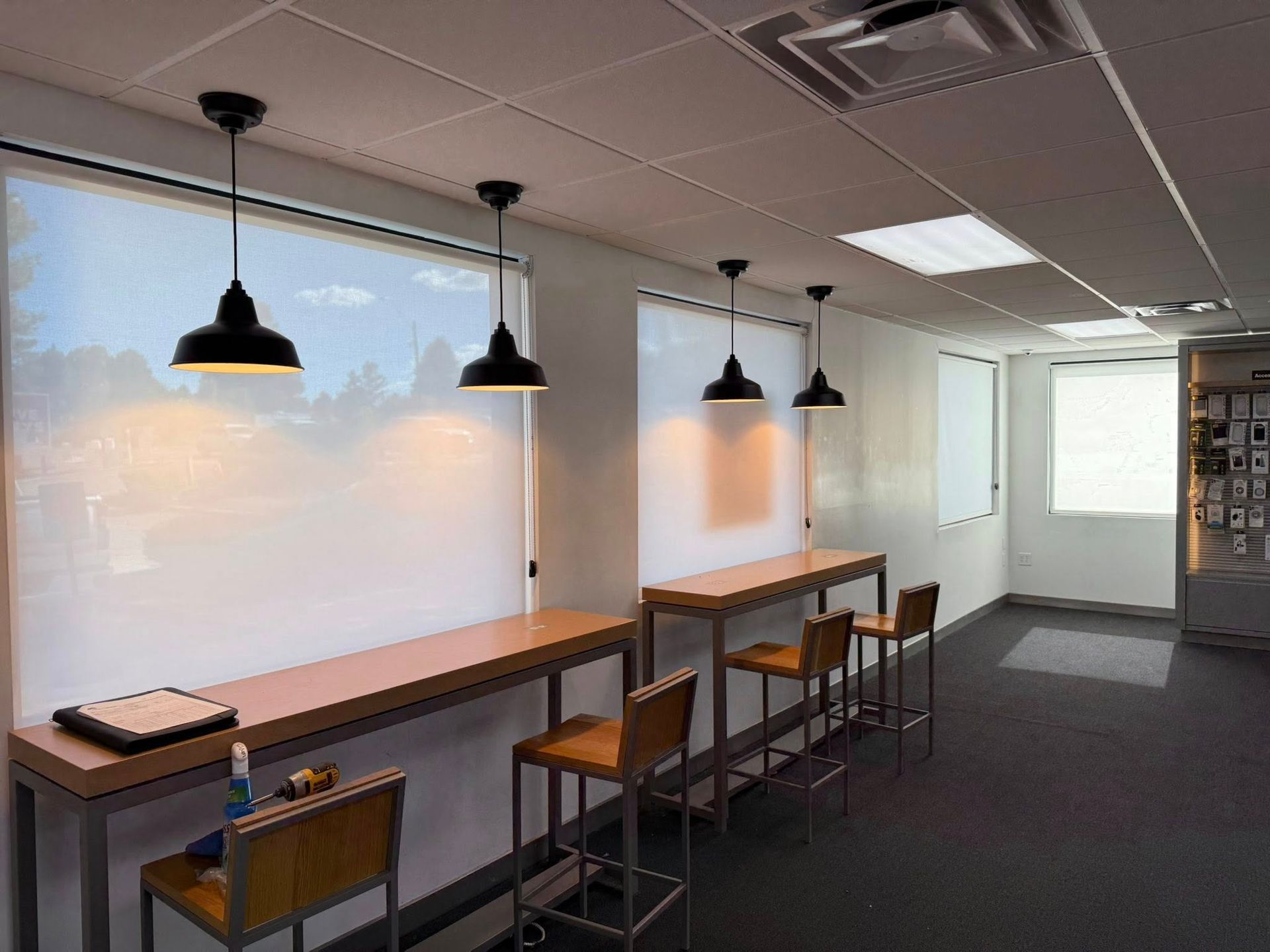 Office break room with high tables, stools, pendant lights, and large window blinds.