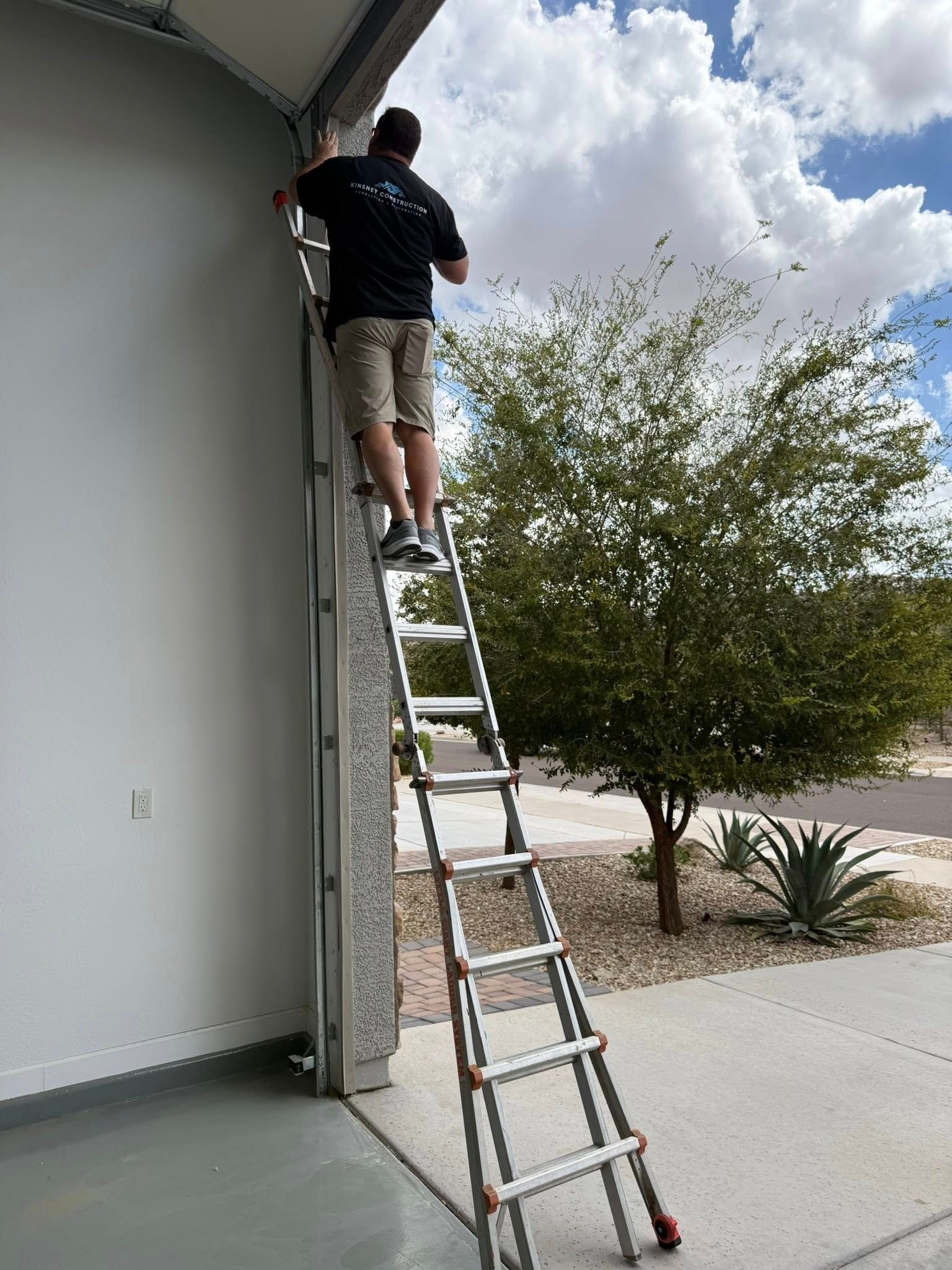 Person on a ladder, adjusting garage door hardware against a light gray wall, outdoors.