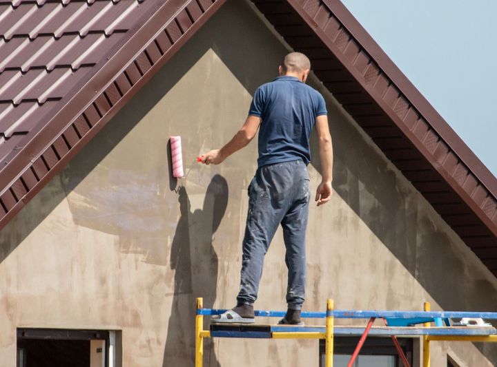 A man is standing on a scaffolding painting the side of a house.