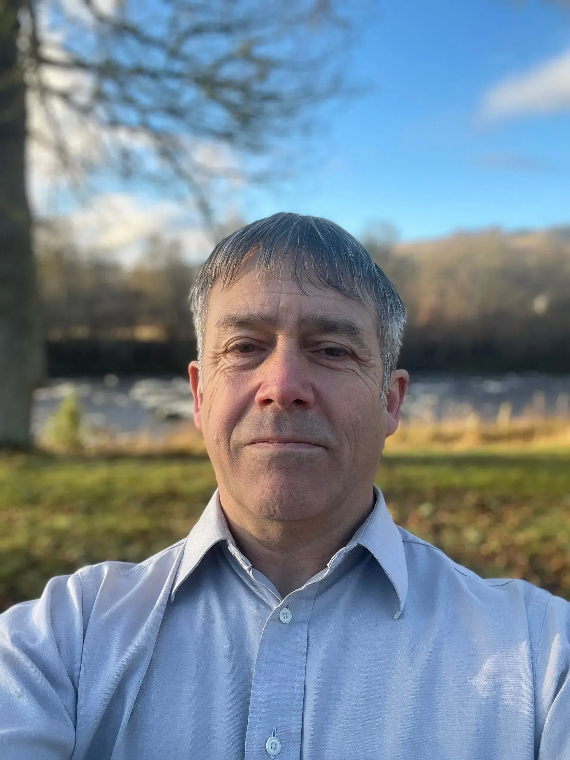 Middle-aged man with graying hair wearing a light blue shirt, looking directly at the camera. Set outdoors, with a blurred background of a river and trees.