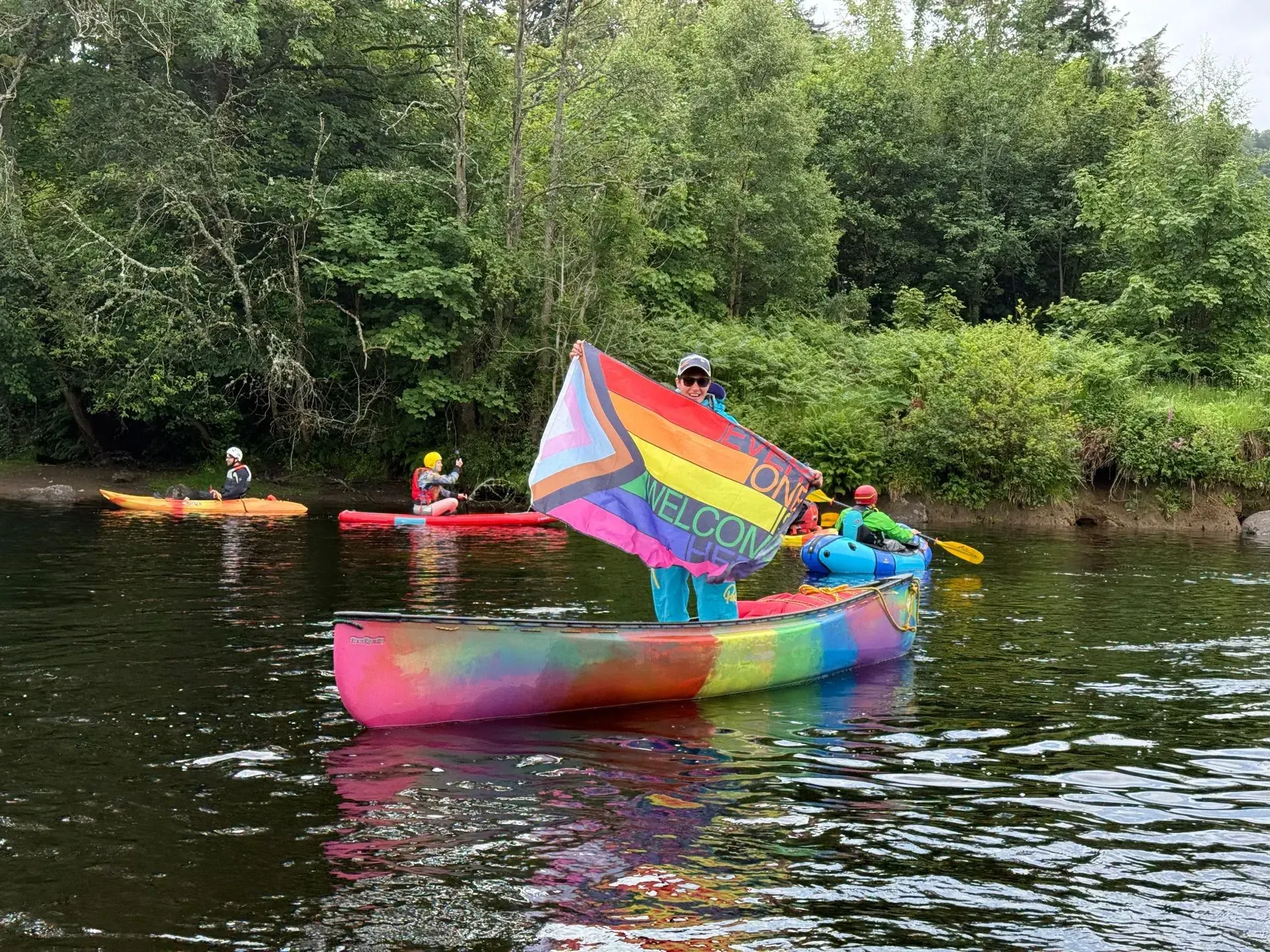 A colorful canoe with a rainbow sail floats on a river, surrounded by kayaks and trees. A person holds the sail.