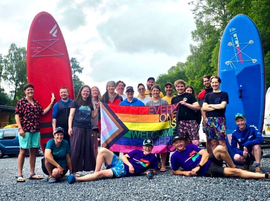 Group of people with a pride flag and paddleboards at a lakeside location. Some people are kneeling or lying down, smiling.