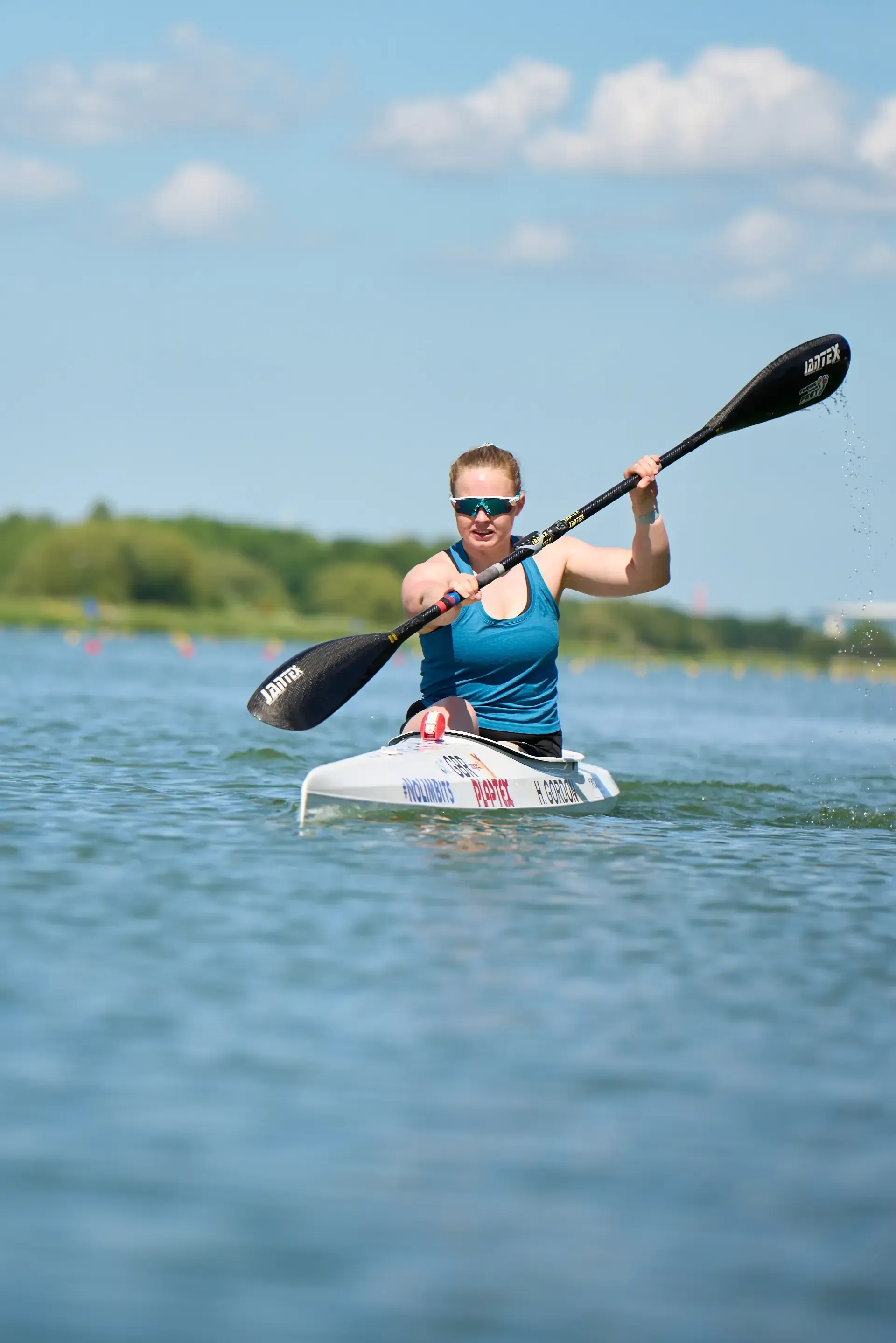 Woman in blue top kayaking on a lake, paddling with a black oar. The sky is blue, and there are trees in the background.