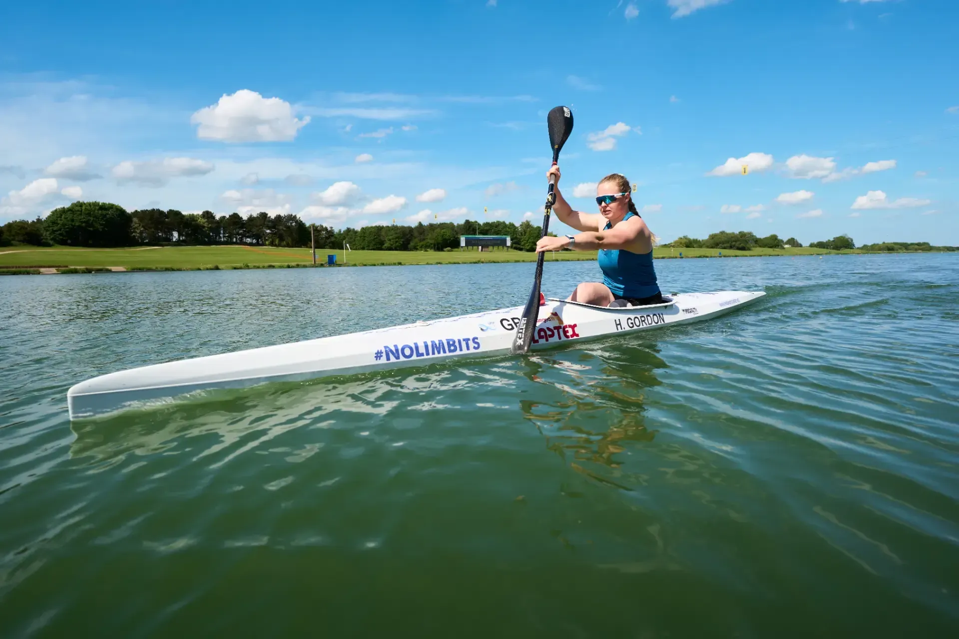 Woman in a kayak on calm water, paddling with a black oar under a blue sky, with green land in the background.