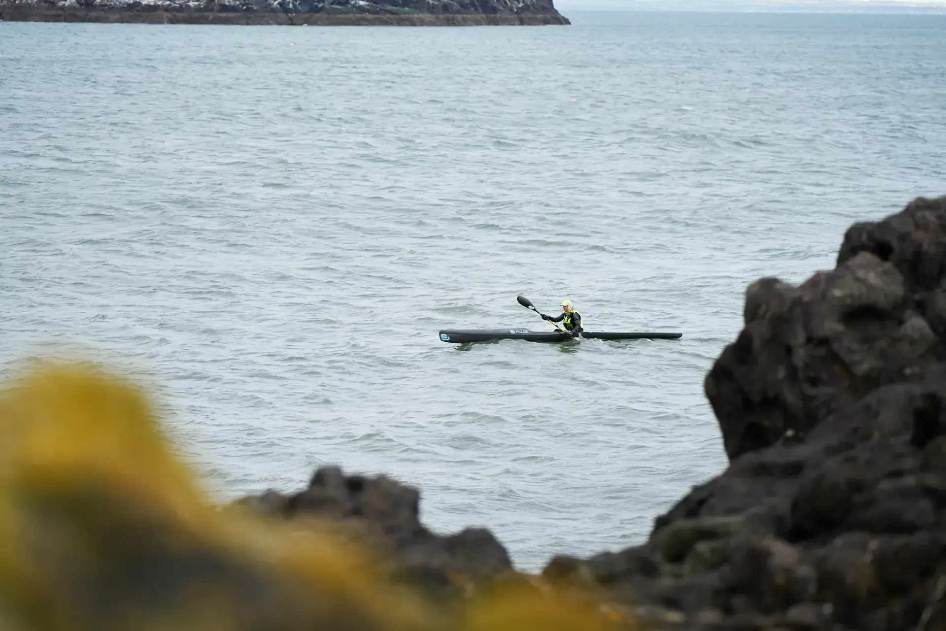 A kayaker in a yellow jacket paddles across a calm sea near a rocky coastline.