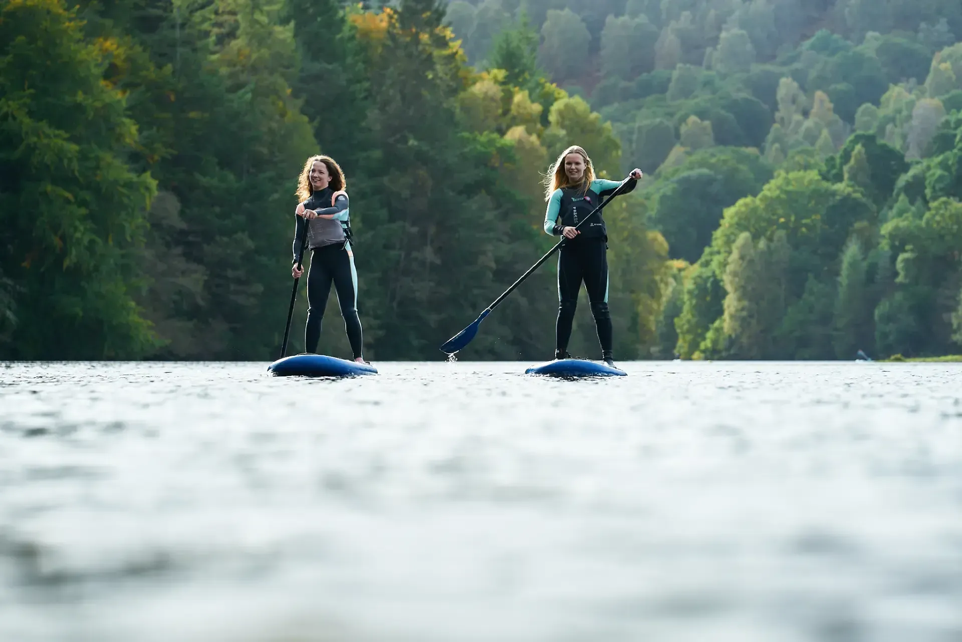 Two women paddleboarding on a lake, wearing wetsuits and smiling, with trees in the background.
