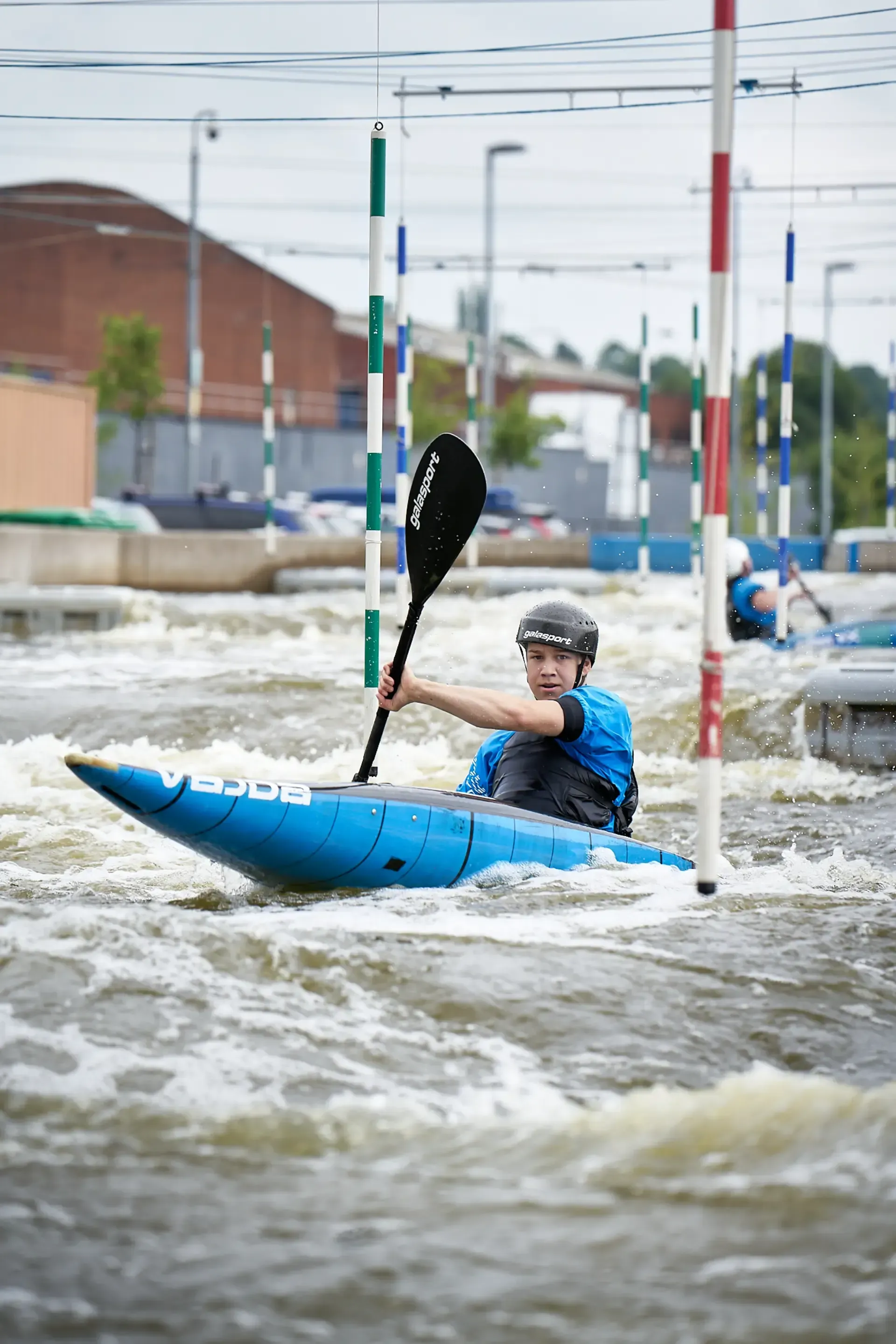 A kayaker in a blue kayak navigates a whitewater course, paddling between vertical poles. The setting is a water channel with buildings visible in the background.