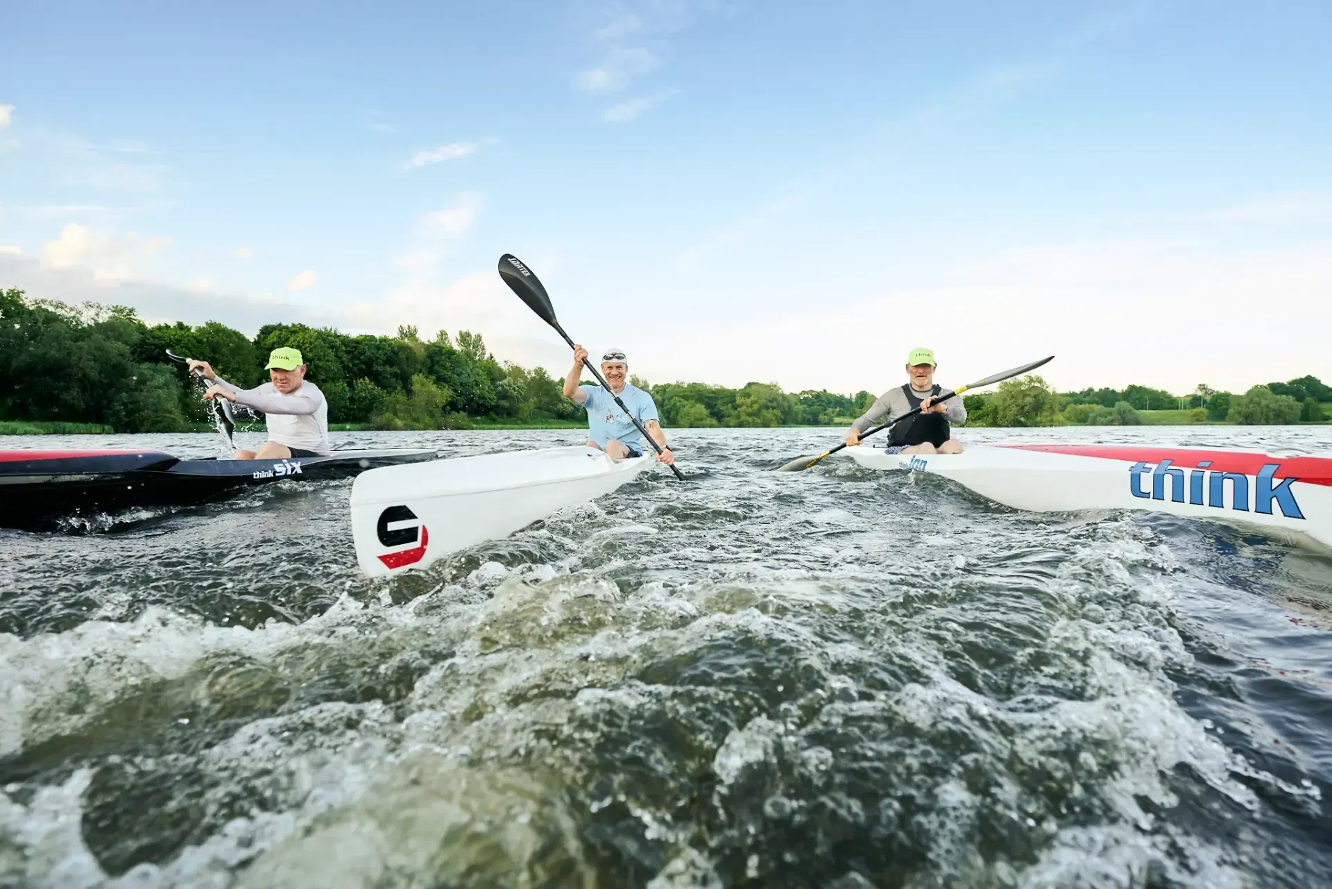 Three kayakers in white kayaks paddle through choppy water under a blue sky.