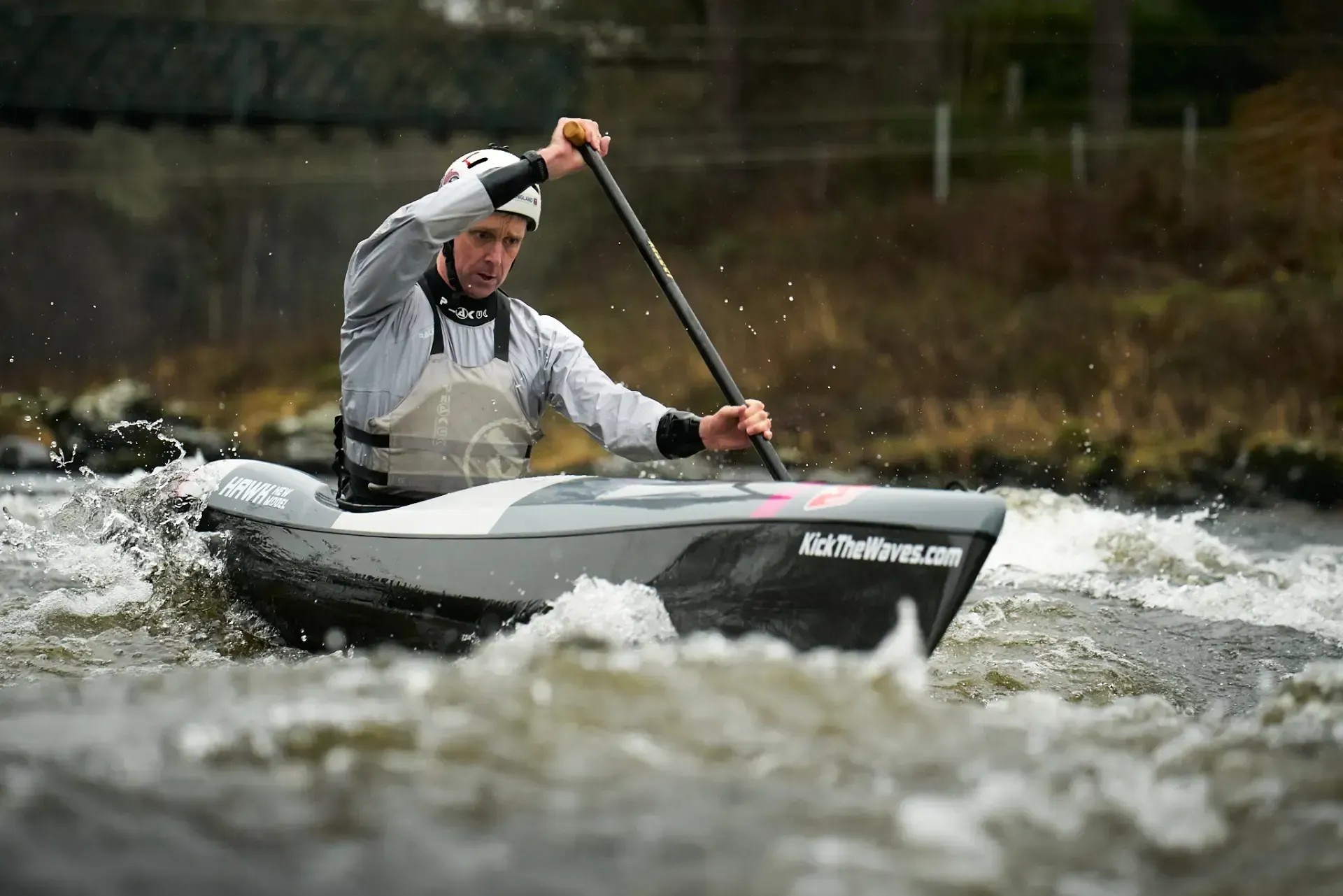 Man in a white helmet and paddling gear navigates a whitewater kayak in a river. Water splashes around the boat in a natural setting.