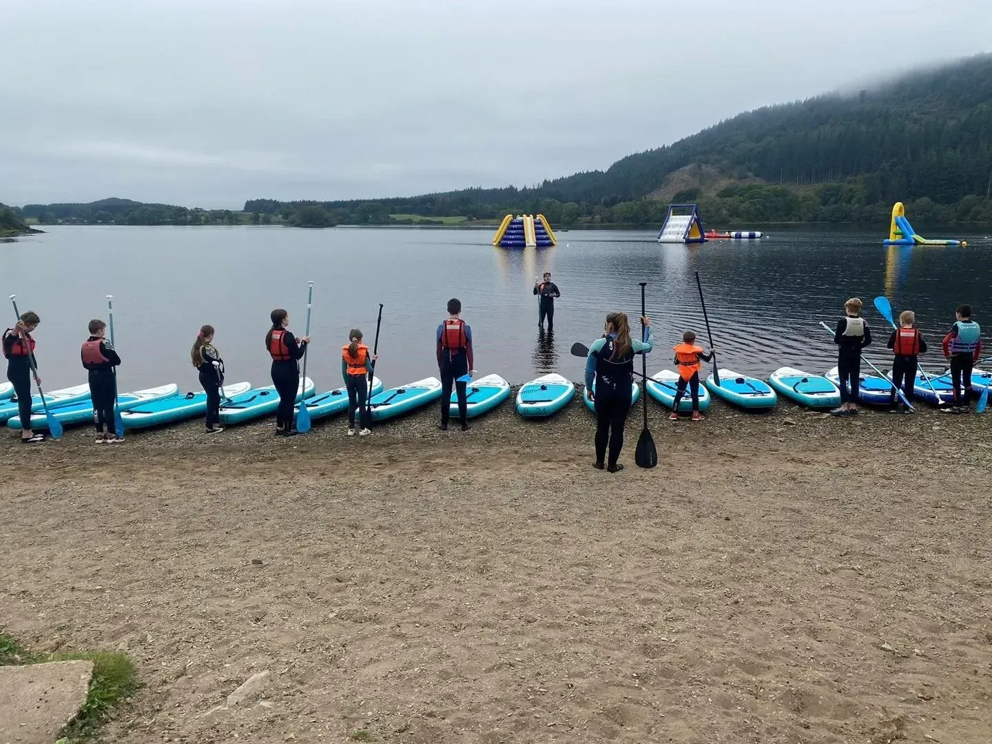 A group of people stand with paddleboards on a lake shore, preparing to paddle. The lake has inflatable water park structures in the background.