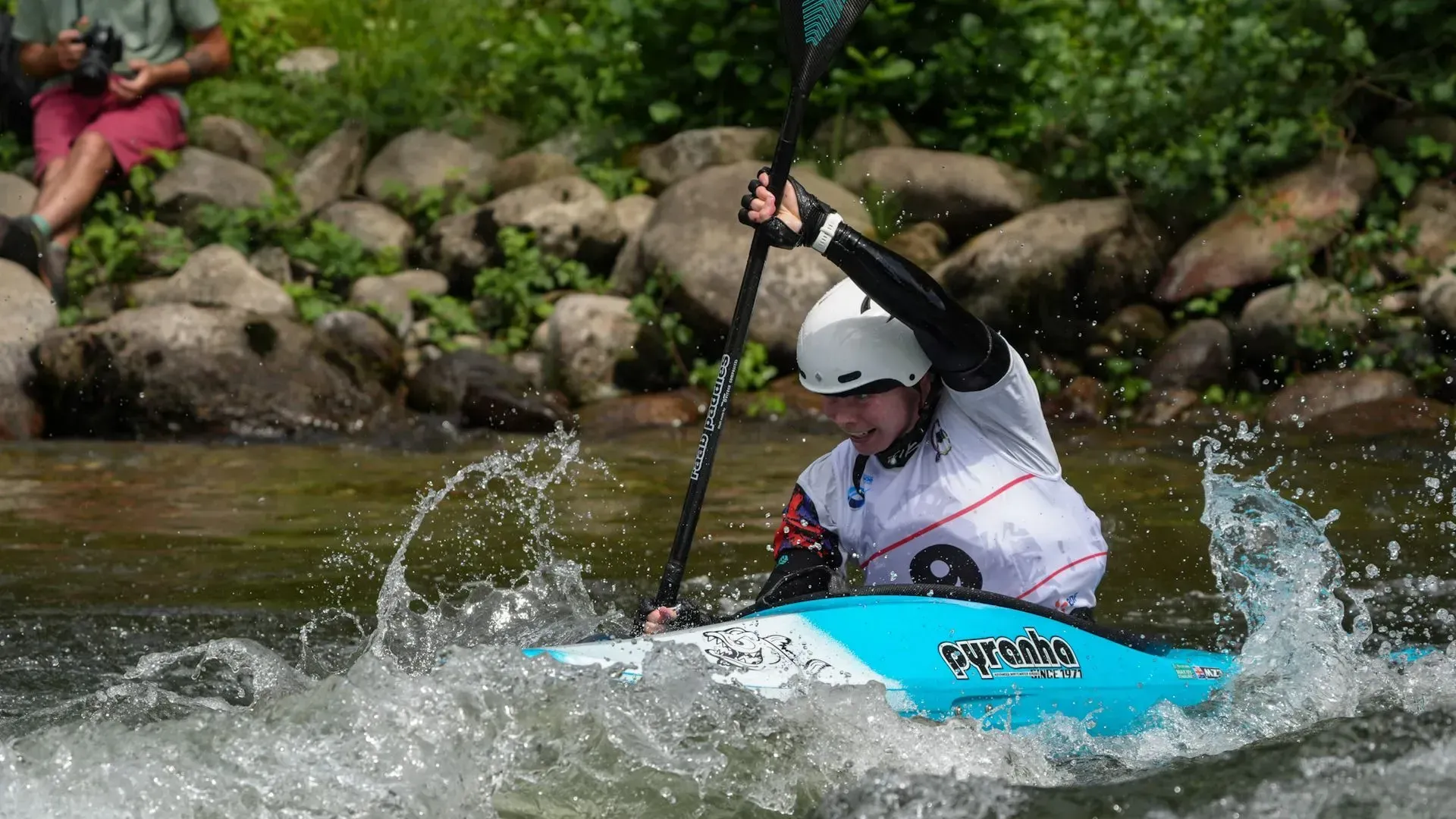 Kayaker in a turquoise boat navigating white water, with rocks and greenery in the background. The kayaker is wearing a helmet and reaching with a paddle.