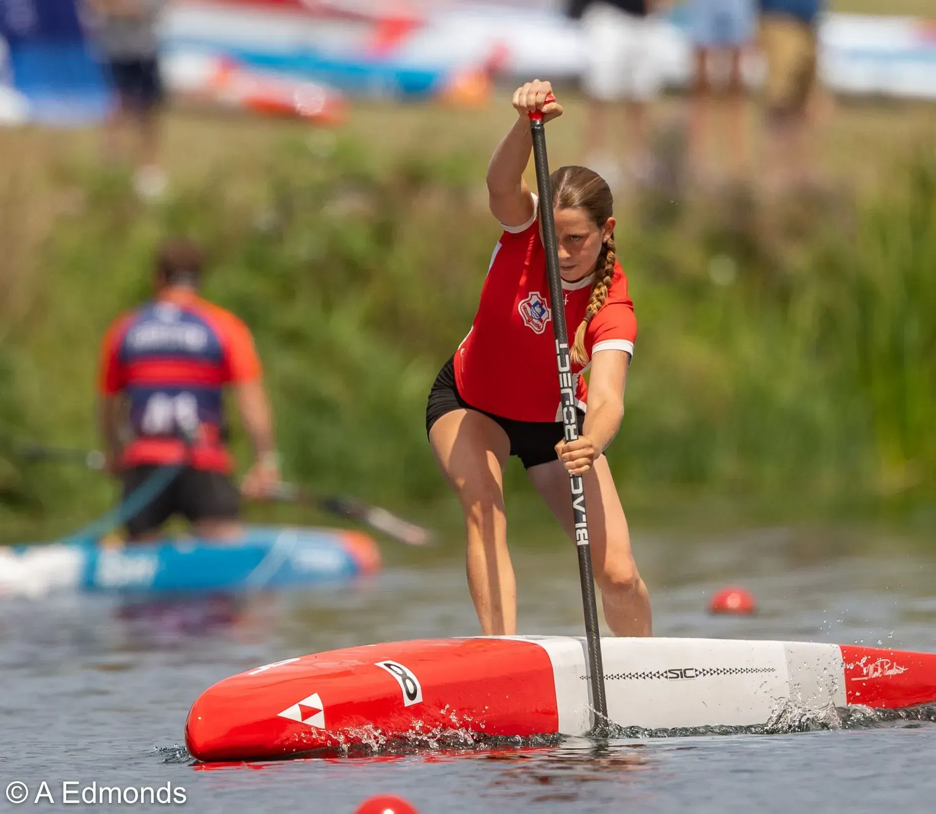 Young woman in red shirt paddles a stand-up paddleboard in a race. She is focused, pushing water with the paddle.