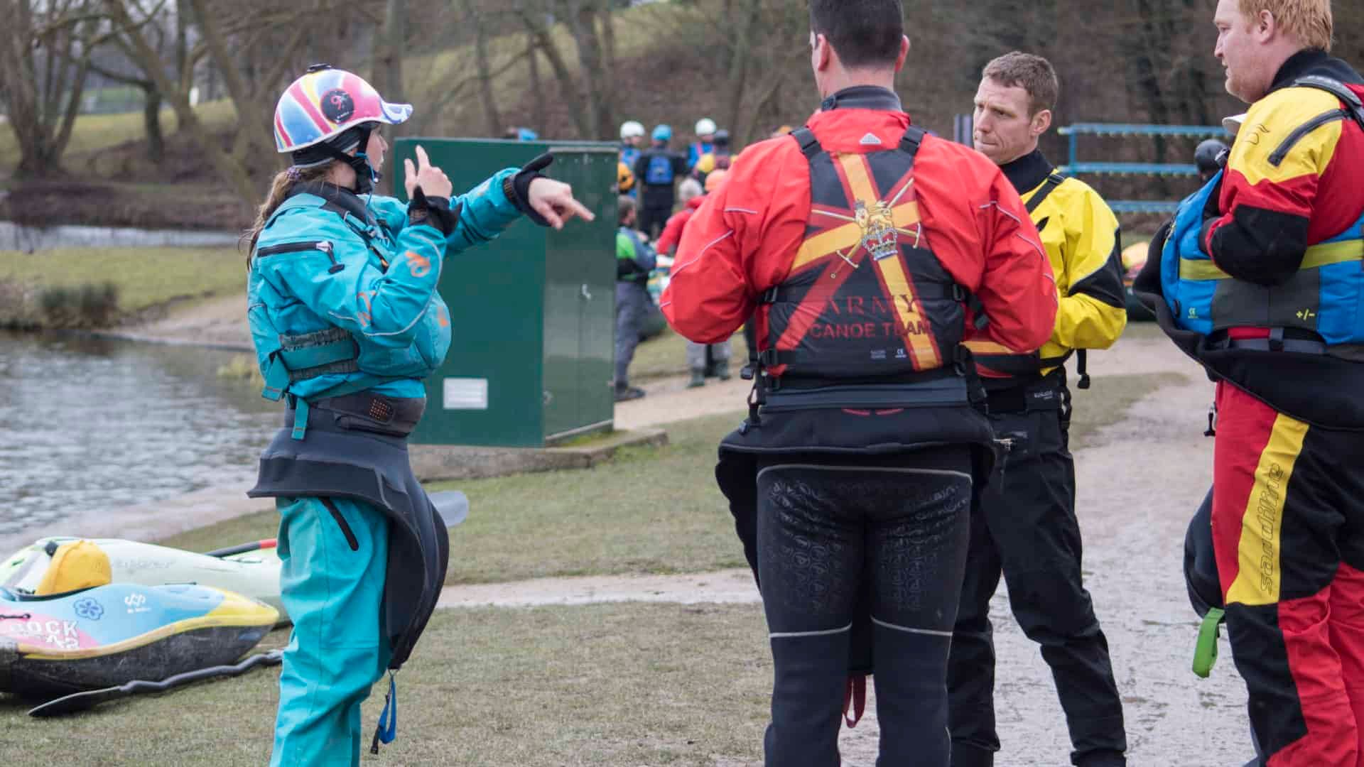 A person in a blue jumpsuit points while talking to a group of people in life vests near a body of water.