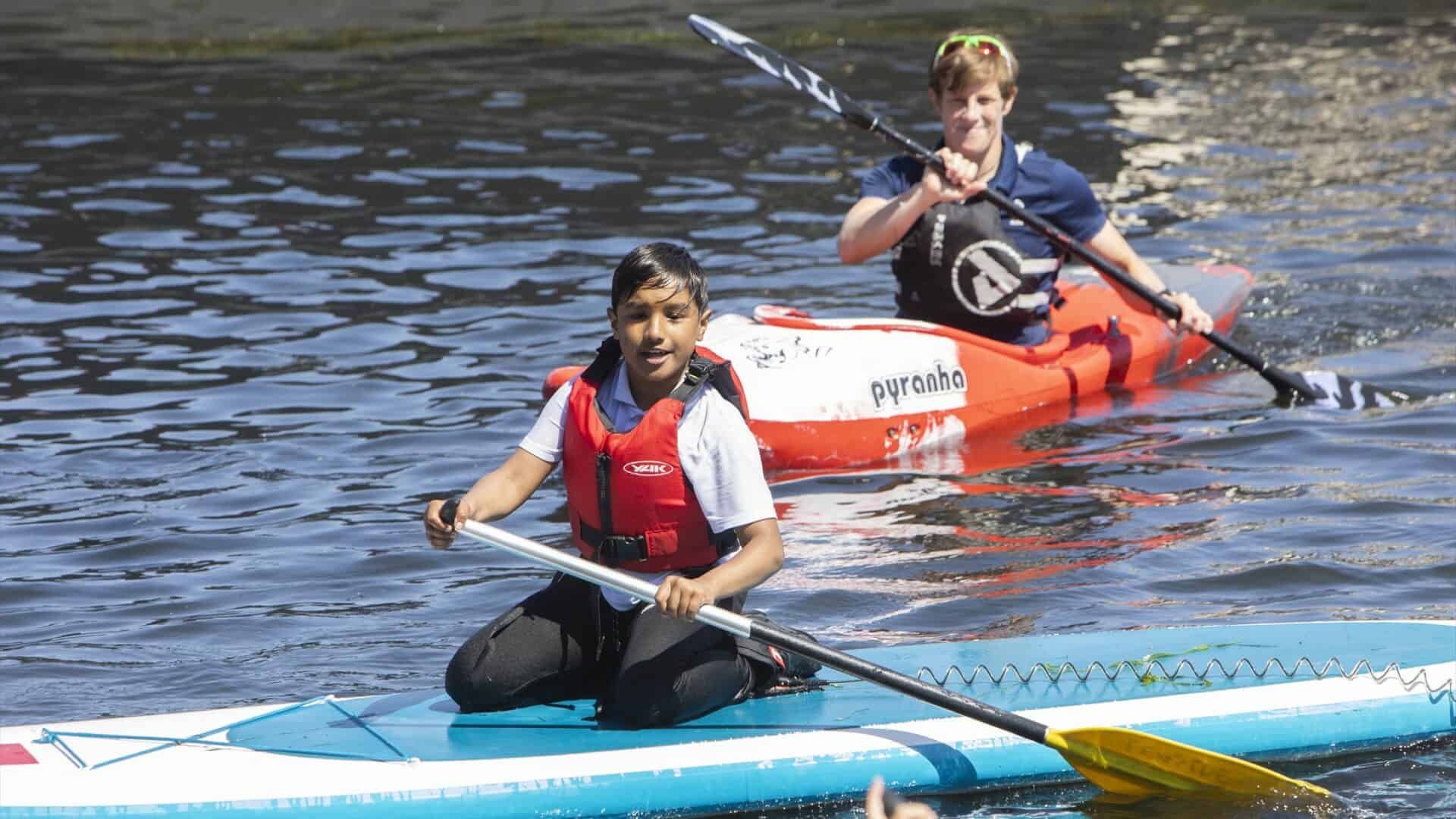 Woman kneeling on a paddleboard, using a paddle. A boy in a kayak paddles behind her on a sunny body of water.