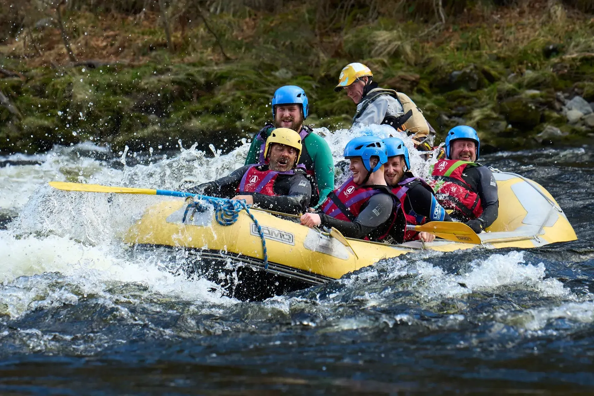 People white water rafting in a yellow inflatable raft on a river, splashing through waves. They wear helmets and life vests.