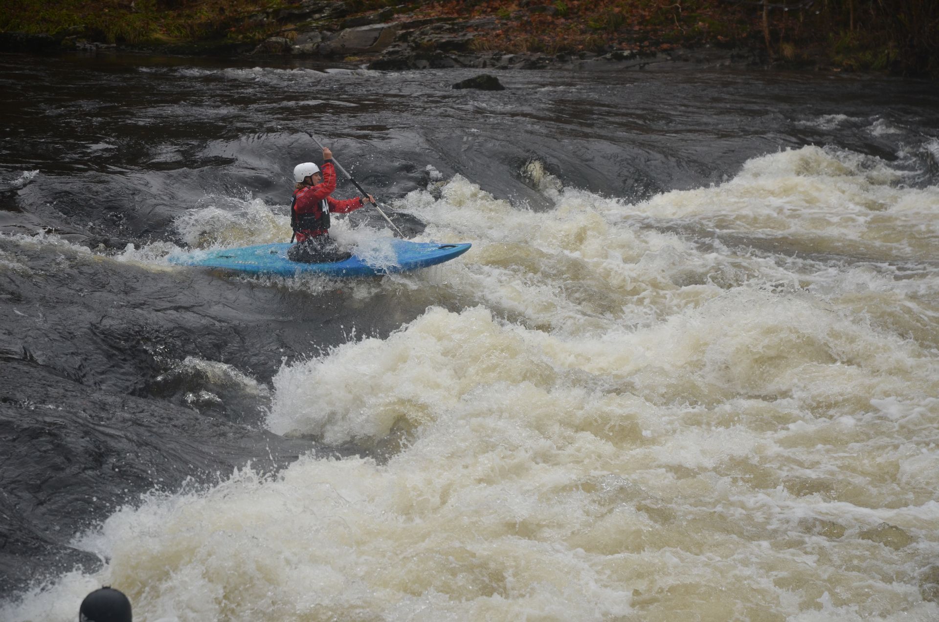 A kayaker in a blue kayak navigates whitewater rapids. They wear a red jacket and paddle, focused in a challenging environment.