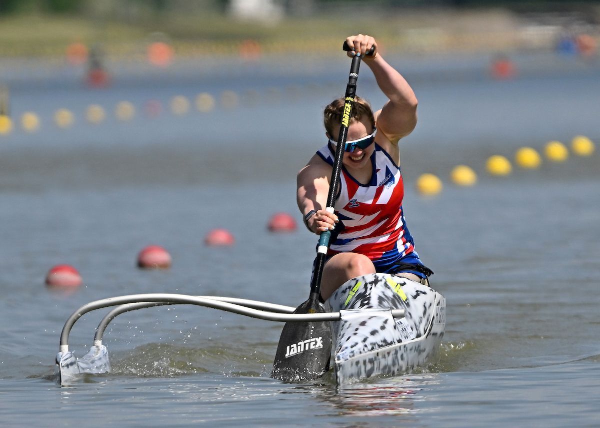 A person in a Union Jack outfit paddles a canoe on a sunny lake, pulling the oar overhead.