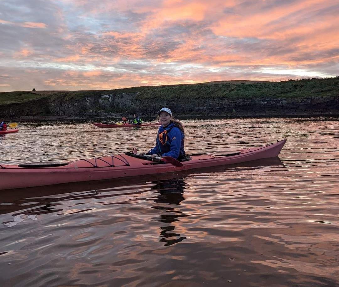 Person kayaking on calm water at sunset. Pink kayak, orange sky, grassy shoreline in the background.