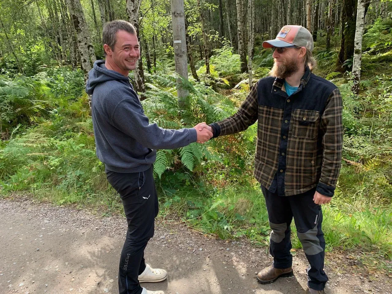 Two men shaking hands outdoors on a forest path. One wears a hoodie and the other a flannel shirt and baseball cap.